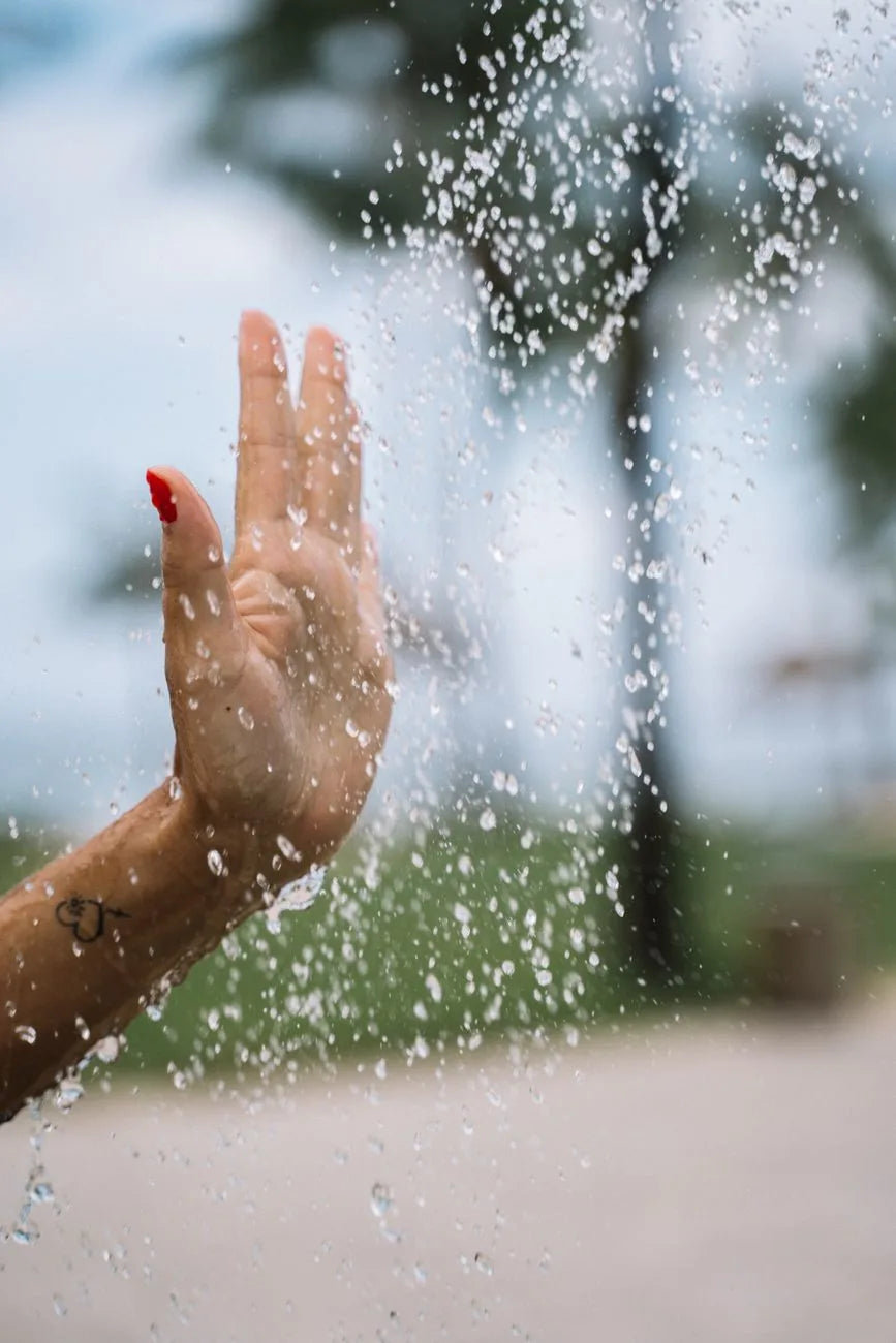 Une main avec des ongles peints en rouge touche des gouttes d'eau tombantes, créant une sensation rafraîchissante en extérieur.