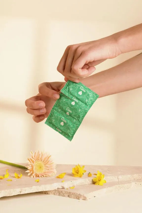 Préservatif écologique en latex naturel, emballage vert, posé à côté de fleurs sur une surface en pierre.